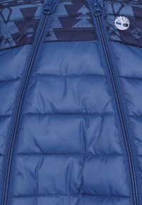 Niebieska pikowana kurtka z dwoma pionowymi zamkami błyskawicznymi oraz wzorem geometrycznych kształtów i logo drzew w okolicy kołnierza.