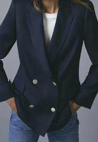 Donna che indossa un blazer doppiopetto blu navy con bottoni argentati sopra una camicia bianca e jeans blu, mani nelle tasche su uno sfondo neutro.