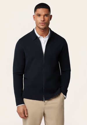 Man draagt een zwarte cardigan met ritssluiting over een wit poloshirt en beige broek, staand tegen een effen lichte achtergrond.