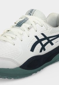 Αθλητικό παπούτσι Asics σε λευκό και σκούρο μπλε με διχτυωτό επάνω μέρος, λευκά κορδόνια και υφή πράσινης σόλας, εμφανίζεται από πλευρική και μπροστινή γωνία.