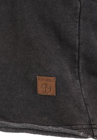 Zwarte stof met een gestructureerd oppervlak, voorzien van een vierkante, bruine leren label met de tekst "KEY LARGO" en een logo in de hoek.