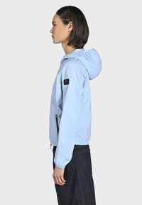 Giacca leggermente azzurra con cappuccio, polsini elasticizzati, tasche con cerniera e un piccolo logo nero sulla manica. Tessuto liscio e leggero.