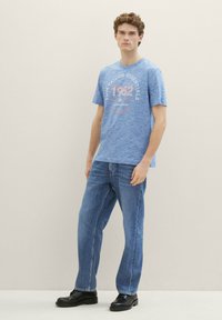 Modra heathered bombažna majica s kratkimi rokavi in okroglim izrezom, ki vsebuje tiskano obliko. V kombinaciji s pravočasnimi denim hlačami in črnimi čevlji.