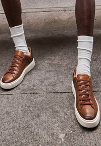 Baskets en cuir marron avec semelles et lacets en caoutchouc blancs, présentant une texture lisse et un design minimaliste. Portées avec des chaussettes blanches.