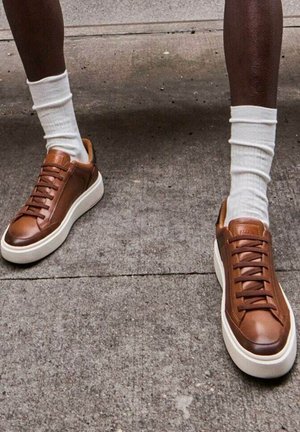 Baskets en cuir marron avec semelles et lacets en caoutchouc blancs, présentant une texture lisse et un design minimaliste. Portées avec des chaussettes blanches.