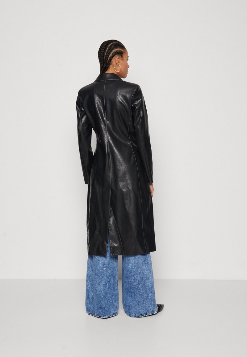 Mujer con cabello trenzado que lleva un abrigo de cuero negro ajustado, jeans anchos azules y zapatos negros, de pie frente a un fondo blanco liso.