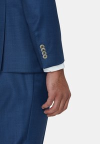 Rękaw niebieskiej marynarki z teksturowanej tkaniny, pięć okrągłych guzików oraz biała mankiet koszuli wystająca spod spodu.