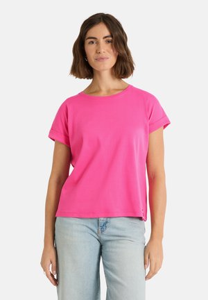 Frau mit schulterlangem braunem Haar, die ein leuchtend rosa kurzärmeliges Oberteil und hellblaue Jeans trägt, vor einem einfarbigen Hintergrund stehend.