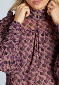 Blusa de manga larga en tejido texturizado, con un patrón floral en púrpura y negro, cuello alto y cierre frontal con botones decorativos.