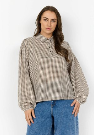 Naine, kellel on seljas beežide ja pruunide ruudulise mustriga pikkade varrukatega pluus kraega ning sinised teksapüksid, seisab ühevärvilise valge tausta ees.