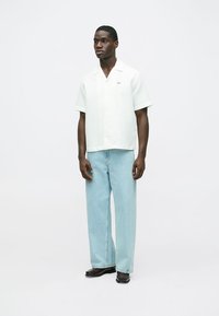 Vit kortärmad skjorta med strukturerad textur, kombinerad med ljusblå löstsittande jeans och svarta skor, stående på en neutral bakgrund.