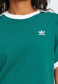 Persona con camiseta verde de Adidas con cuello blanco y logo en el lado izquierdo del pecho, mostrando solo la parte superior del torso y el cuello.