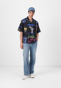 Cămașă neagră cu mânecă scurtă, cu imprimeuri grafice colorate, asortată cu blugi deschiși la culoare și adidași albi. Modelul poartă o șapcă bleu.