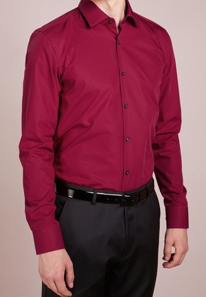 Homme portant une chemise bordeaux à manches longues boutonnée, rentrée dans un pantalon noir avec une ceinture noire brillante, debout devant un fond uni.