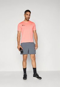 Herridrottsoutfit med en lysande rosa kortärmad tröja och grå shorts, med svarta sportstrumpor och skor, håller en vattenflaska.