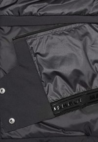 Schwarze isolierte Jacke mit einem Innenfutter, das eine mit Reißverschluss verschließbare Tasche und matte Beschläge aufweist. Glatte Textur mit einem elastischen Akzent in Markenoptik.
