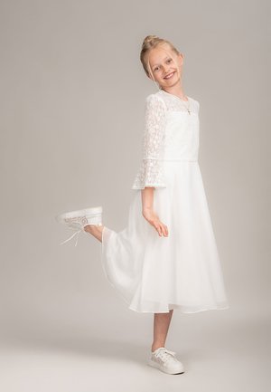 Weißes Spitzenkleid mit dreiviertellangen Ärmeln und gerafften Manschetten, kombiniert mit weißen Turnschuhen. Das Kleid verfügt über einen fließenden Rock und dezente Details.