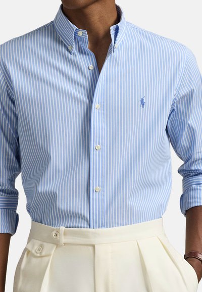 Chemise à rayures bleues et blanches à boutonnières, avec des manches longues, présentant un petit logo brodé et un col classique, associée à un pantalon de couleur crème.