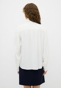 Blouse blanche à manches longues en tissu lisse, avec un dos plissé et un ourlet arrondi, associée à une jupe bleu marine.