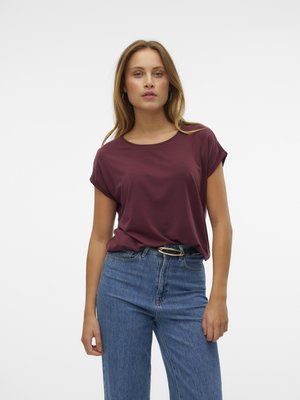 Frau mit langen blonden Haaren, die ein kurzärmliges bordeauxrotes Oberteil trägt, das in eine hoch taillierte blaue Jeans mit schwarzem Gürtel und goldener Schnalle gesteckt ist.