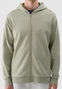 Felpa verde chiaro con zip realizzata in materiale morbido, dotata di una cerniera frontale, due tasche laterali e polsini e orlo a coste.