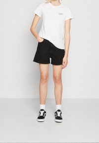 Camiseta de algodón blanca con gráfico de "Levi's", combinada con pantalones cortos de mezclilla negros. El modelo lleva calcetines blancos y zapatillas negras. Conjunto simple y casual.
