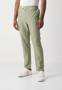 Man wearing light green drawstring linen pants and white sneakers, standing against a plain white background.