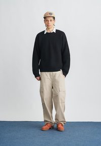 Pulover negru tricotat, pantaloni cargo bej cu buzunare laterale și pantofi portocalii. Este purtat o șapcă bej, cu un logo. Fundal simplu.