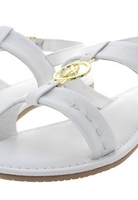 Bílé kožené sandály mají překřížené popruhy, kulatý zlatý akcent a plochou gumovou podrážku se šitými detaily.