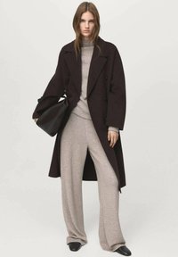 Lange donkerbruine jas over een lichtgrijze trui en broek met wijde pijpen. Soepele stof met een losse pasvorm. Zwarte tas en schoenen.