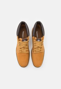 Botas de ante marrón con un collar de cuero negro, cordones planos y plantillas con marca. Presentan ojales de acero y detalles de costura en contraste.