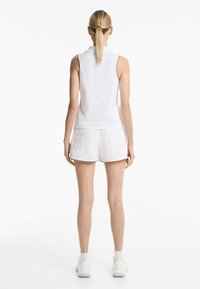 Une femme aux longs cheveux blonds se tient de dos, vêtue d'un haut sans manches blanc et d'un short, avec un fond blanc uni.