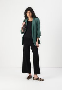 Blazer verde sopra una maglietta nera, abbinato a pantaloni larghi neri e ballerine con stampa leopardo. Il blazer ha le maniche arrotolate e una vestibilità comoda.