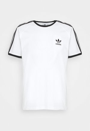 Wit katoenen T-shirt met een zwarte ronde hals en zwarte biezen aan de mouwen. Heeft drie zwarte strepen langs elke schouder en een klein logo op de borst.