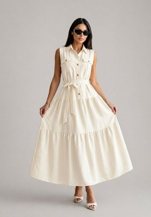 Femme portant une robe longue beige sans manches boutonnée avec ceinture, tenant les côtés de la jupe, lunettes de soleil et talons ouverts sur fond gris.
