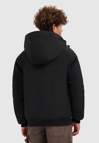 Een persoon met krullend haar, gekleed in een zwarte hoodie en bruine cargo broek, staat met hun rug naar de camera op een grijze achtergrond.