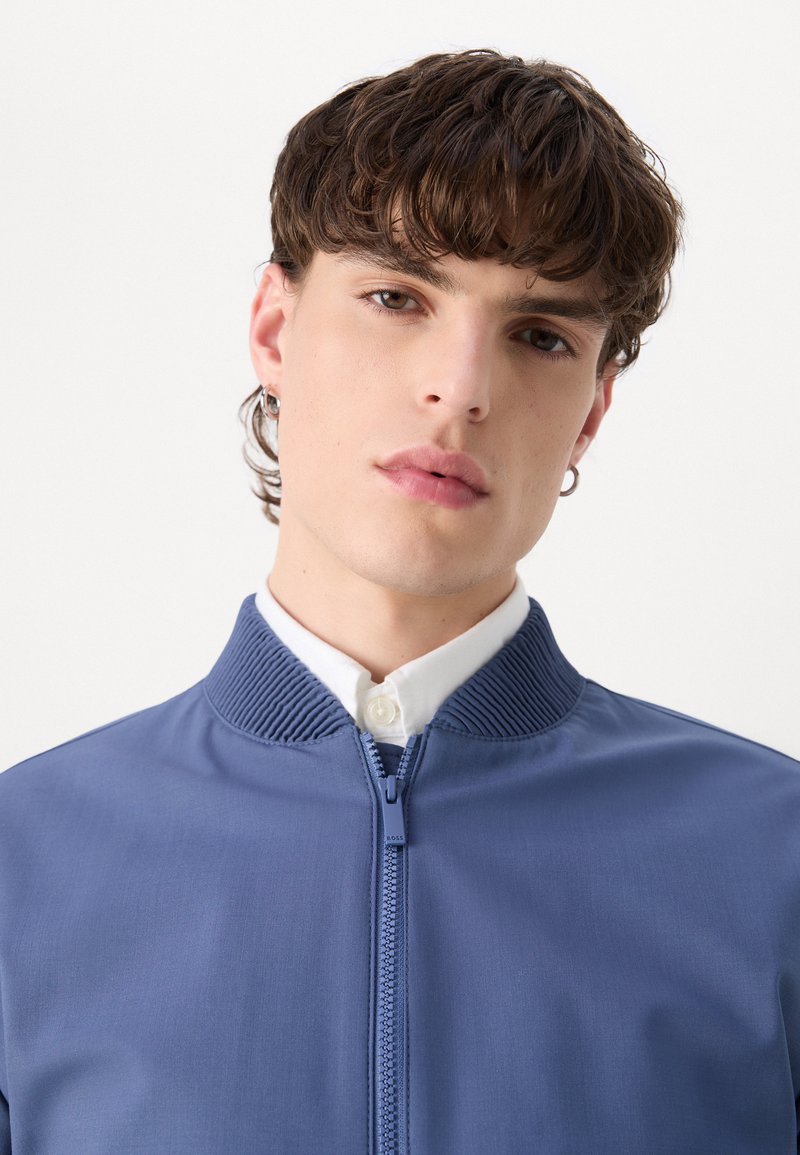 Veste bleue zippée avec un col côtelé, texture lisse et une chemise blanche à boutons en dessous. Design simple et coupe ajustée.