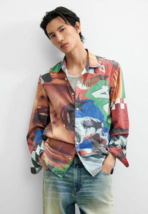 Joven con cabello corto y oscuro lleva una camisa de botones colorida con patrones abstractos y vaqueros azules descoloridos, manos en los bolsillos, fondo neutro.