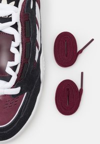 Bordowo-czarny tenisówka z białymi sznurowadłami, obok dwóch starannie zwiniętych bordowych sznurowadeł na białym tle.