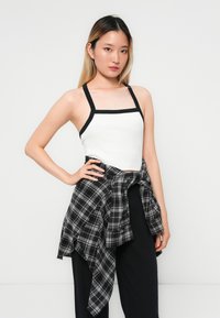 Hvit ribbete tanktop med svart kant, kombinert med en knyttet svart og grå rutete flanellskjorte. Modellen har på seg løse svarte bukser.