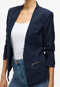 Veste de blazer marine à coupe slim, devant ouvert et manches trois-quarts. Comprend une poche zippée sur le côté gauche et une texture de tissu lisse.
