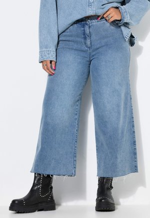 Person, die hellblaue Jeans mit weitem Bein und rohen Säumen, eine hellblaue Jeansjacke und schwarze, genietete Lederstiefel trägt.