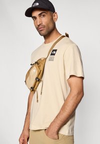 Μπεζ πουκάμισο με κοντά μανίκια και μαύρο λογότυπο, συνδυασμένο με καφέ τσάντα χιαστί με φερμουάρ και ρυθμιζόμενους ιμάντες, φορεμένη στον αριστερό ώμο.