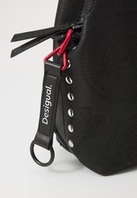 Zwarte faux leren tas met gegraveerde patronen, zilveren studs en een rode hardwareaccent. Inclusief een gemerkte schouderband met een lus.