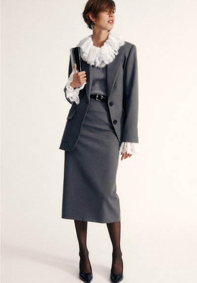 Sivina obleka z ruffled belim ovratnikom, vzorčastimi rokavi in ujemajočo se krilom. Na nogah ima črne pete in drži ročno torbico.