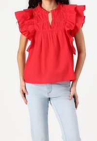 Blouse rouge à manches volantes, avec un col en forme de goutte et une broderie délicate, associée à un jean bleu clair. Texture de tissu lisse.