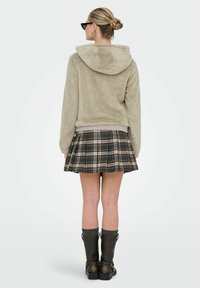 Faux-Fur-Hoodie in hellem Beige, mit plüschiger Textur und einer breiten Kapuze. Kombiniert mit einem gefältelten Karorock in dunklen Tönen und kniehohen schwarzen Stiefeln.