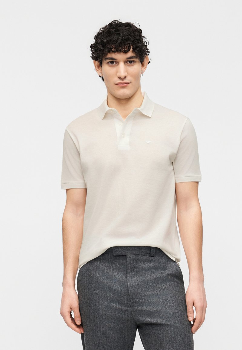 Άνδρας με σγουρά σκούρα μαλλιά, φορώντας μπεζ κο ντομάνικη πόλο μπλούζα και σκούρα γκρι παντελόνια, στέκεται μπροστά σε λευκό φόντο.