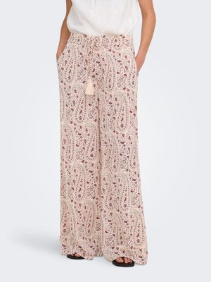 Vrouw draagt beige wijde broek met hoge taille en rood paisley- en bloemenprint, gecombineerd met een witte mouwloze top en zwarte sandalen.