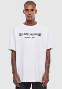 Balta per didelė t-shirt marškiniai su juodais grafikos tekstais: „gyvenimas yra kasdienis triūsas“ ir „UPSCALE ST.“, trumpomis rankovėmis, pagaminta iš medvilnės.
